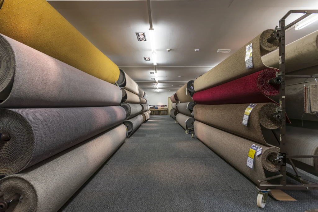 A wide showroom aisle with rows of heavy carpet rolls stacked horizontally on industrial shelving.